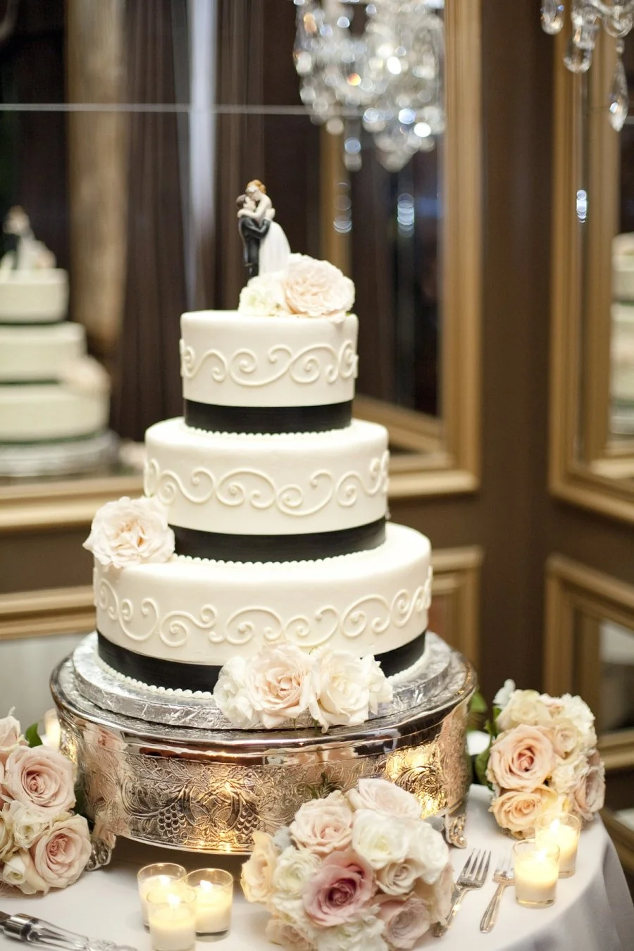 Three-tier wedding cake decorated with white and black fondant, floral accents, and a bride and groom cake topper, surrounded by bouquets of roses and small candles, in an elegant reception setting.