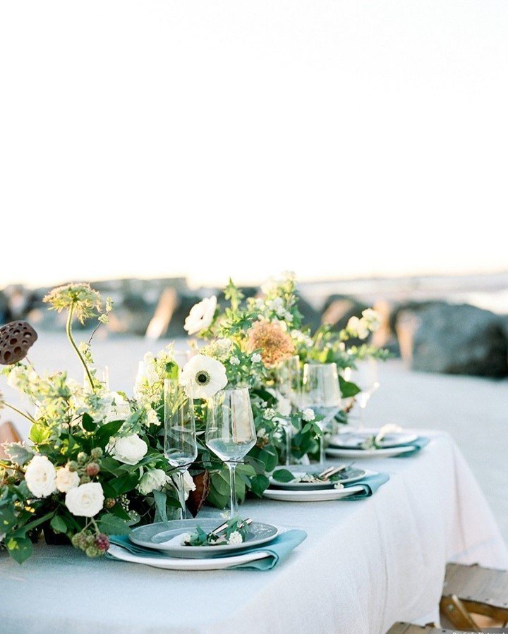 A decorated outdoor dining table with white tablecloth, floral centerpiece, plates, glasses, and napkins, set against a beach or rocky waterfront background.