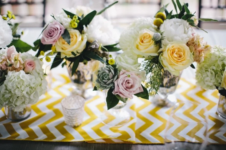 Flower arrangements with roses, succulents, and other flowers in small vases on a yellow and white chevron tablecloth.