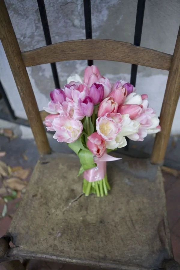 A bouquet of pink and white tulips with green stems and leaves, tied with a pink ribbon, resting on an old wooden chair.