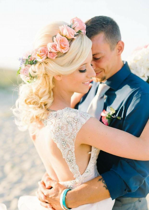 Bride and groom embrace on a beach during sunset, the bride wearing a floral crown and lace dress, the groom in a navy shirt and gray tie.