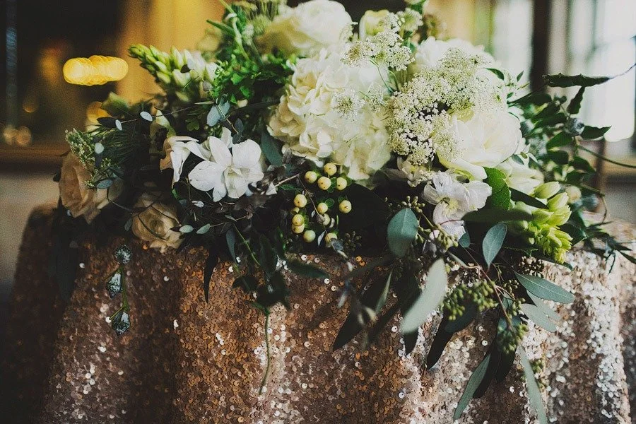 A floral arrangement of white flowers and green foliage on a sparkling, sequined tablecloth.