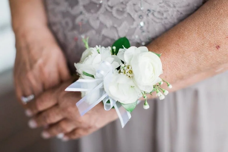 Close-up of a woman's arm with a white floral corsage and bow. She is wearing a lace dress and has her hands clasped.