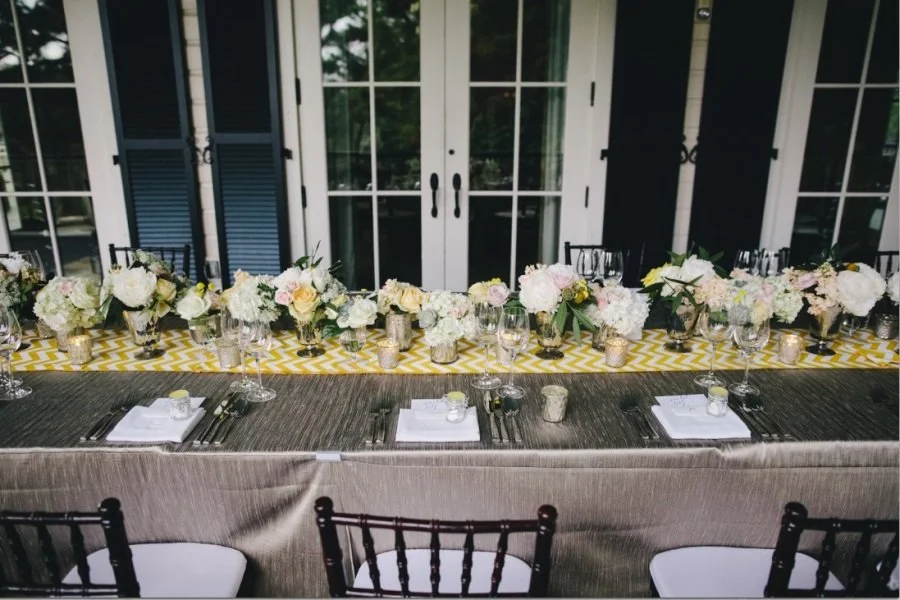 Long dining table decorated with floral centerpieces, candles, and glassware, set for a formal event or celebration inside a house with glass doors and windows.