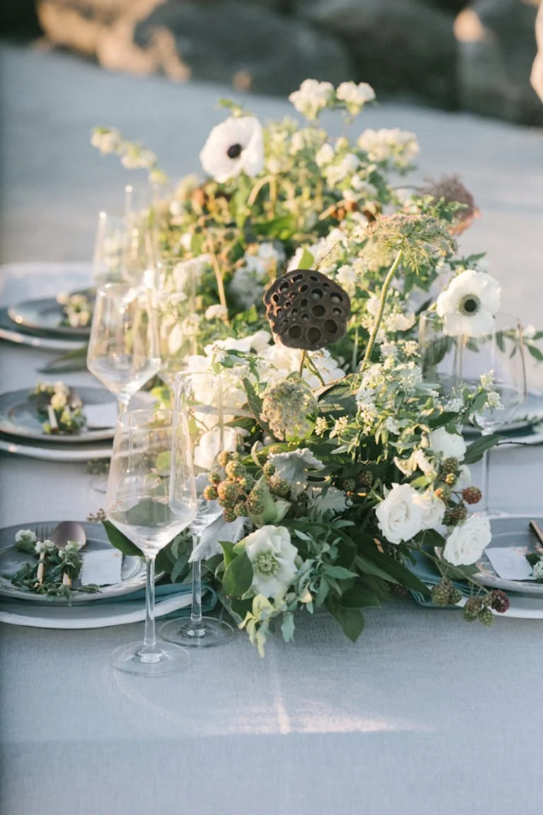 Elegant outdoor table setting with a floral centerpiece, white flowers, greenery, and decorative dark seed pods, with wine glasses and plates arranged on a light-colored tablecloth.