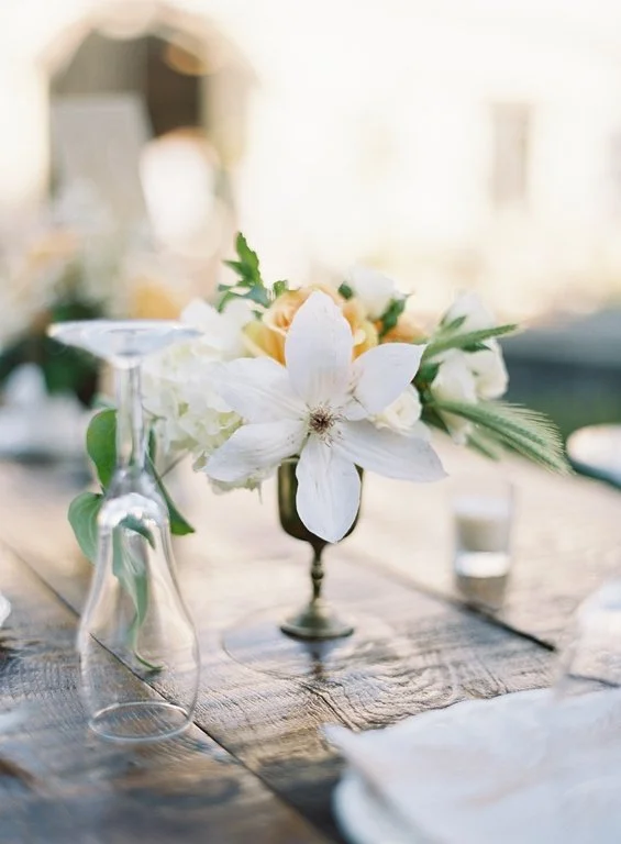 A floral centerpiece featuring white lilies and light-colored flowers on a wooden table, with glassware and a candle in the background.