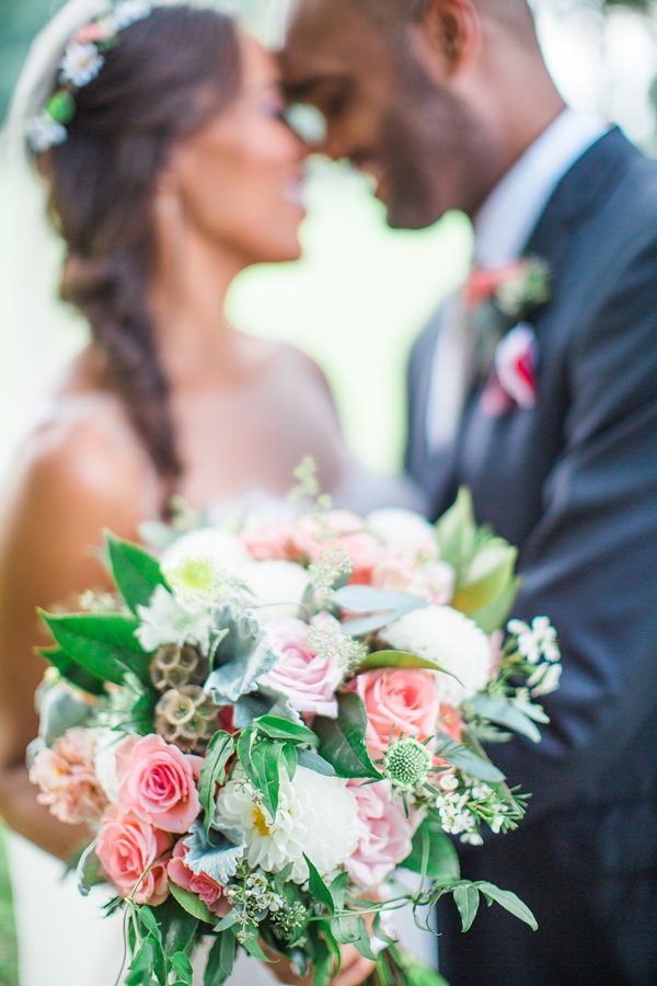 A bride and groom on their wedding day holding a bouquet of pink, white, and green flowers with a blurred background.