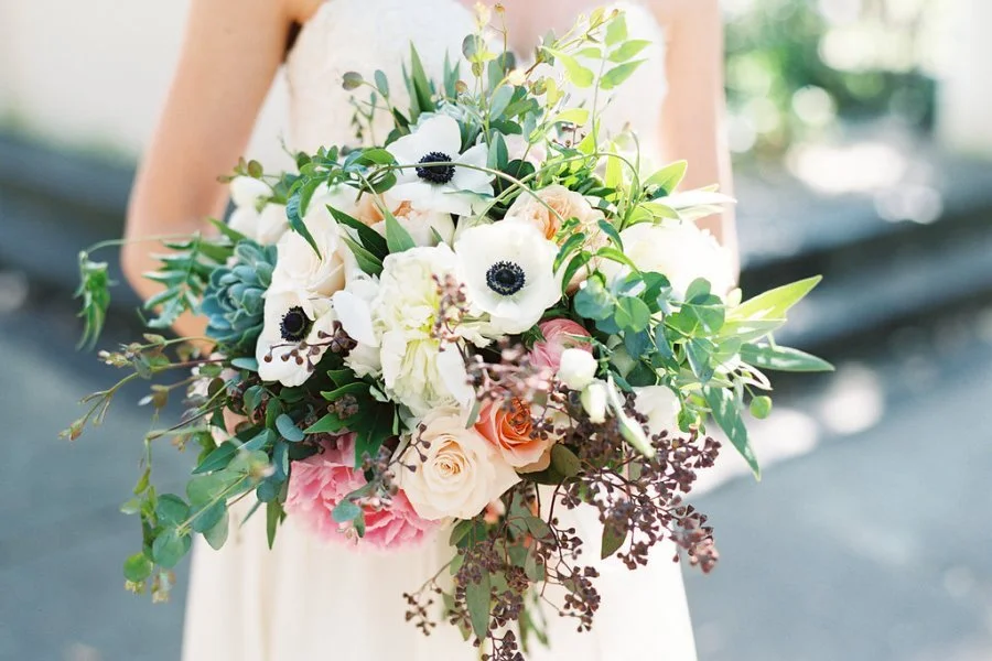 A bride holding a large bouquet of flowers, including white anemones, roses, and various greenery, in an outdoor setting with blurred background.