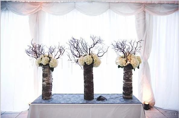 Three tall vases with white hydrangea flowers and dried branches on a table during a wedding or event decor setup.
