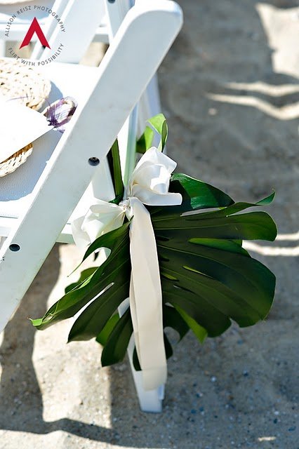 White chair decorated with a large white bow and green tropical leaves tied to its leg, outdoors on sandy ground.