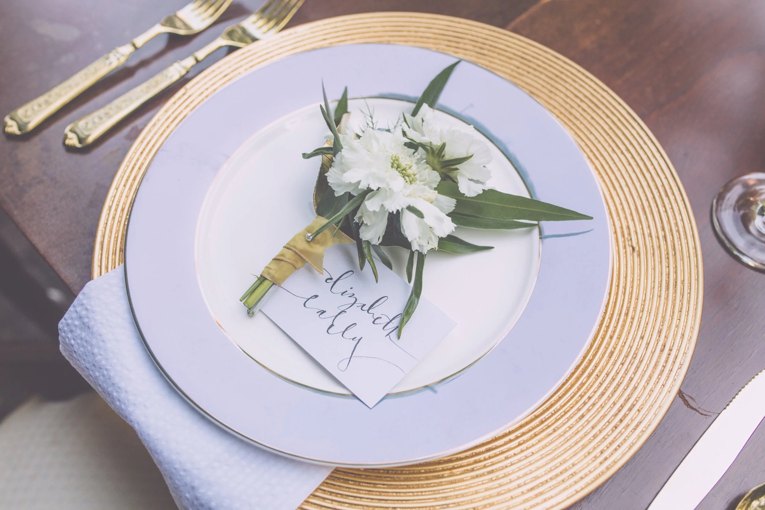 A decorative place setting featuring a white and gold plate, a white napkin, and gold utensils; a small bouquet of white flowers with green leaves and a handwritten place card reading 'Elizabeth Callay'