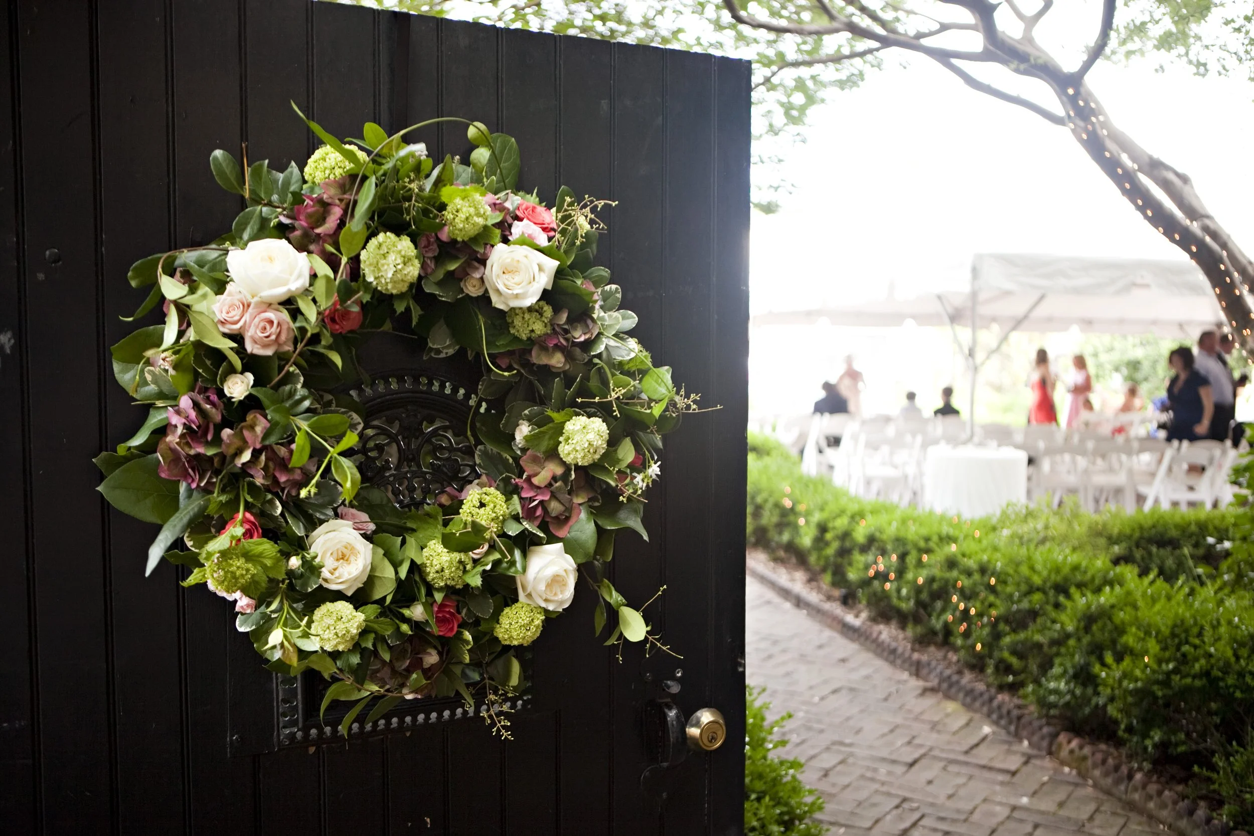 A decorative floral wreath made of white, pink, and green flowers and greenery hung on a black door at an outdoor event with tables and people in the background.