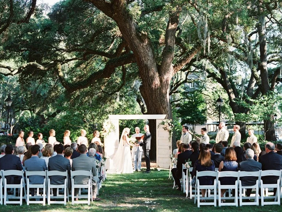 Outdoor wedding ceremony under a large oak tree with brides and grooms exchanging vows, surrounded by guests seated on white chairs.