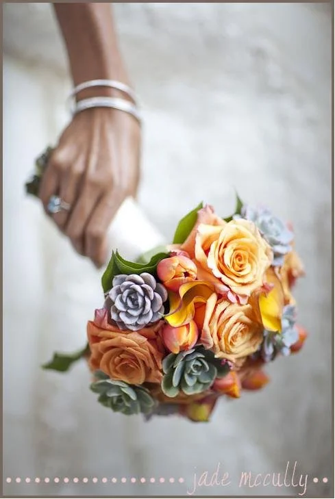 A woman holding a bouquet of multicolored roses and succulents, with the focus on the bouquet and her hand, which has jewelry and a wedding ring.