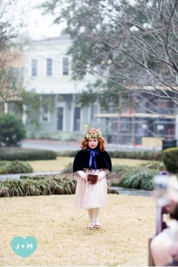 A young girl with curly red hair wearing a floral crown, a black cape, and a light-colored dress, standing on a grassy area with trees and houses in the background.