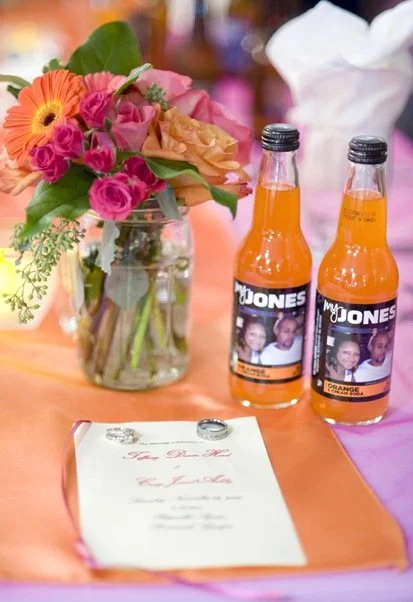 Vase with pink, orange, and purple flowers and two bottles with custom labels on a table with a peach-colored tablecloth.