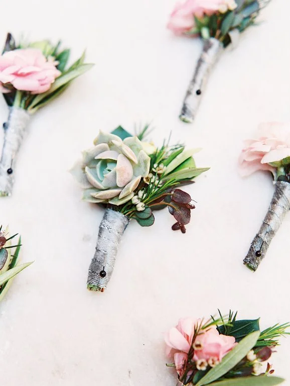 Multiple boutonnieres with pink and white flowers and succulents, wrapped with gray ribbon, scattered on a white surface.