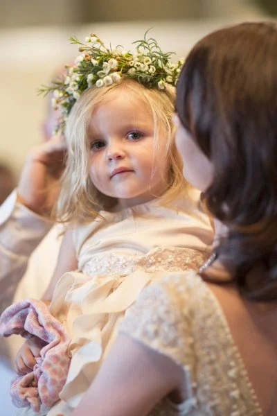 A young girl wearing a flower crown looks at an adult woman who is holding her, during a special occasion.