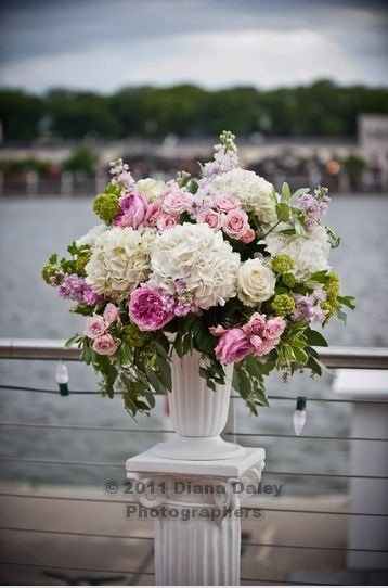 A large bouquet of pink, white, and purple flowers in a white vase on a pedestal near a body of water.