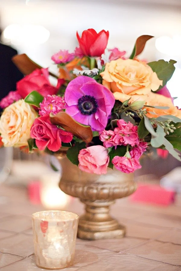 A bouquet of colorful flowers including pink roses, purple anemone, peach roses, and a red tulip in a silver vase on a wooden table with a small candle holder in the foreground.