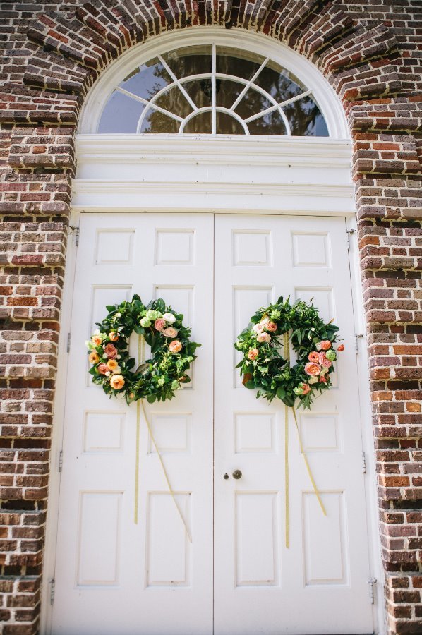 White double doors with decorative wreaths hanging on each door, set in a red brick building with an arched window above.