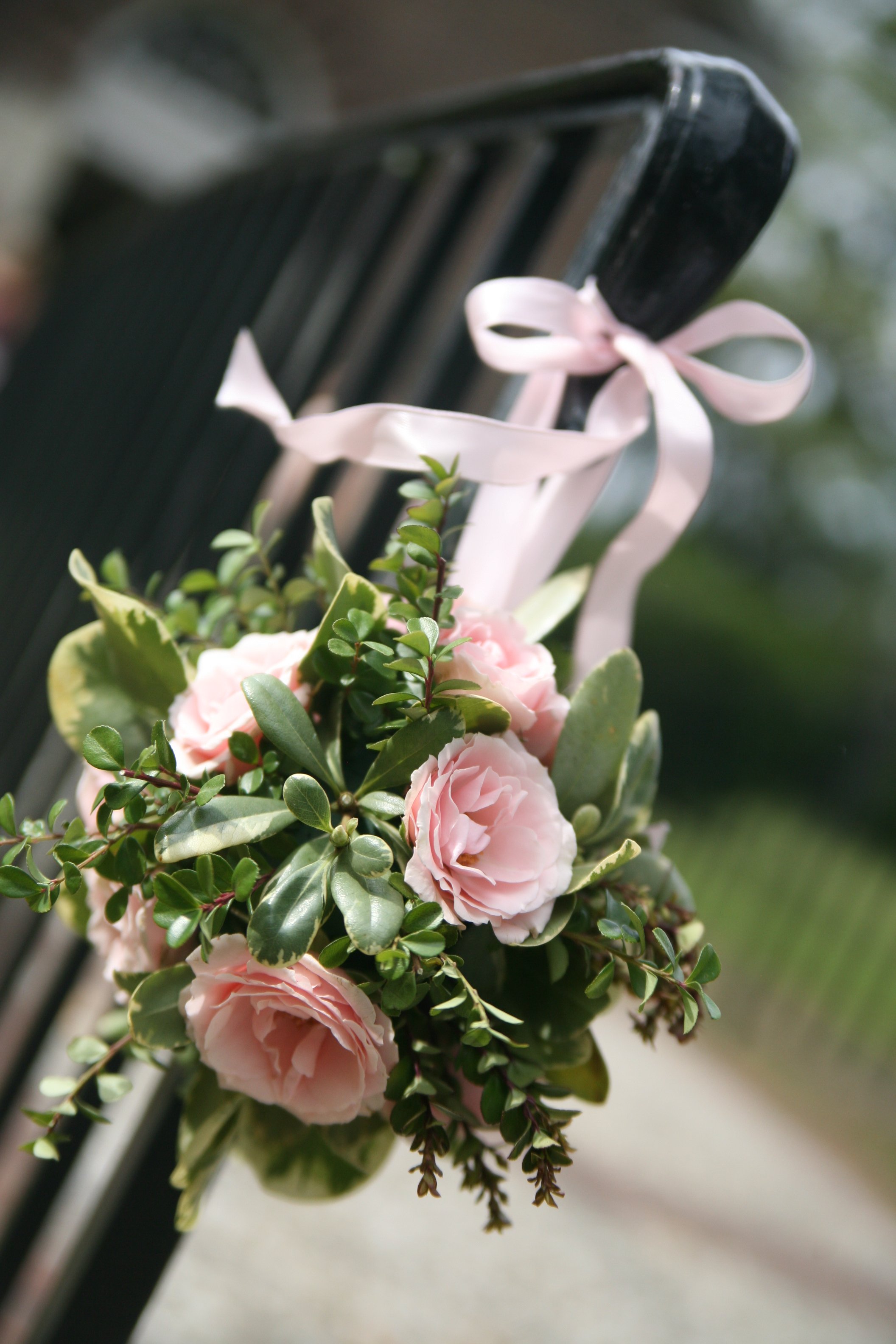 A floral arrangement with pink roses and green leaves hanging attached with a light pink ribbon tied in a bow, on a black metal structure outdoors.