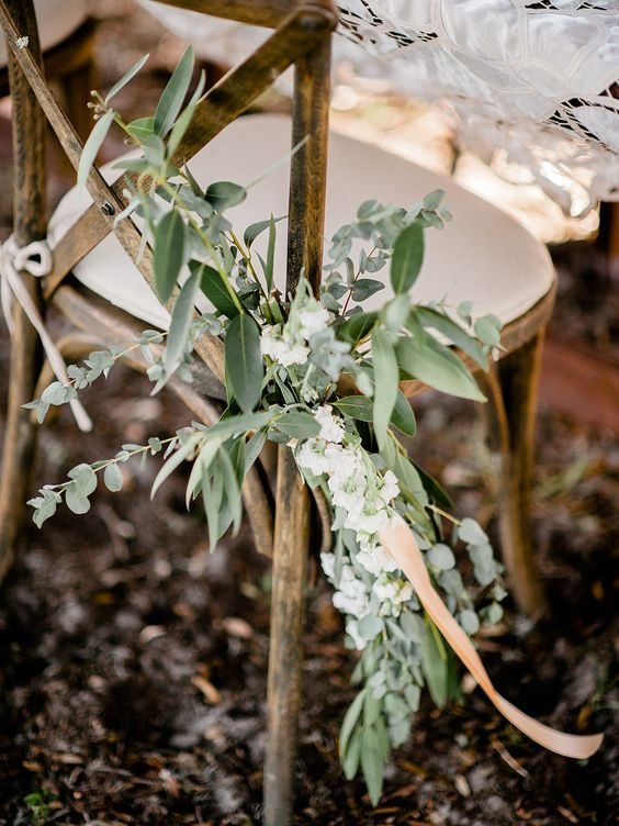 Close-up of a wooden chair with a floral arrangement hanging on its side, featuring green leaves and white flowers, tied with a light pink ribbon, outdoors with soil at the base.