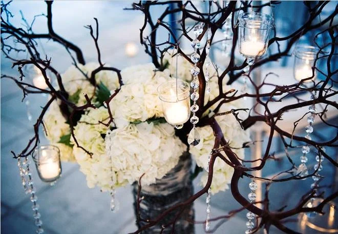 A floral arrangement with white hydrangeas inside a glass vase, surrounded by dark, leafless branches decorated with hanging candle lanterns and strings of clear beads, creating an elegant centerpiece.