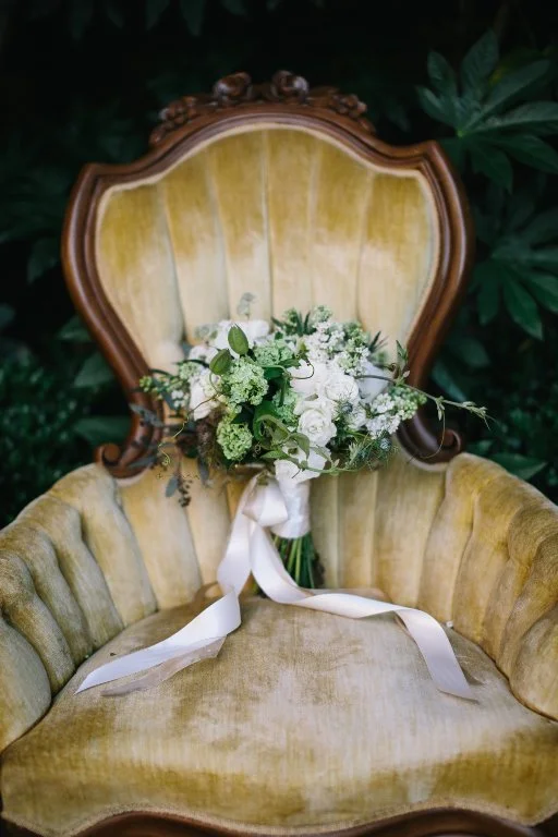 A vintage yellow upholstered armchair with ornate wooden frame, decorated with a white floral bouquet tied with a white ribbon.