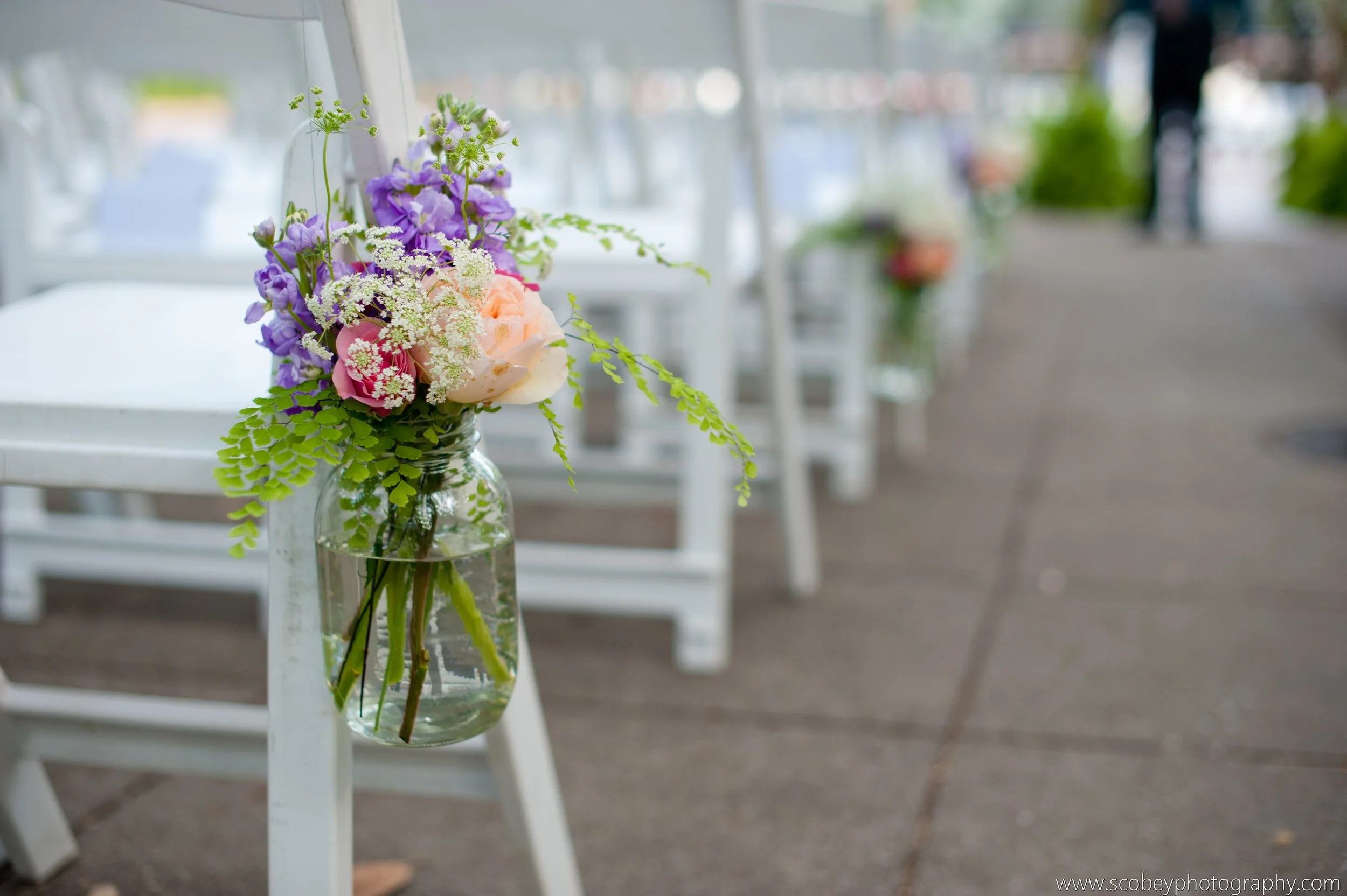 A small glass jar with a bouquet of pink, purple, and white flowers, and green leaves, hanging on the side of a white chair on an outdoor patio or walkway.