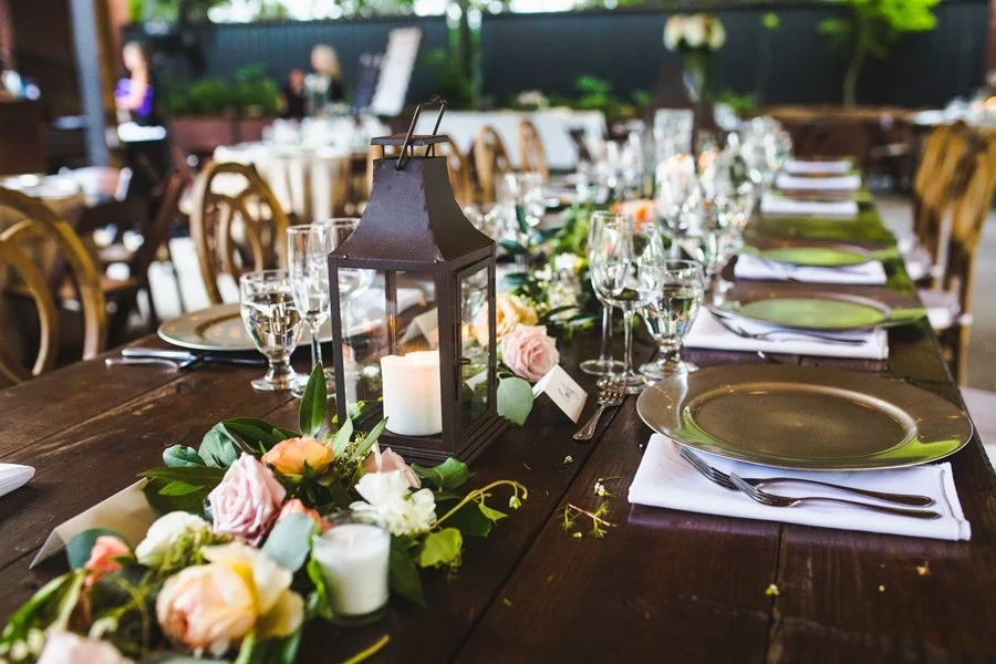 A long wooden table decorated for an outdoor event with a black lantern candle holder, flowers, glasses, plates, silverware, and white napkins.