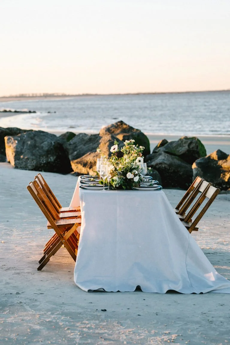 A table set for a meal on a beach with rocks and ocean in the background, featuring a white tablecloth, floral centerpiece, and multiple place settings.