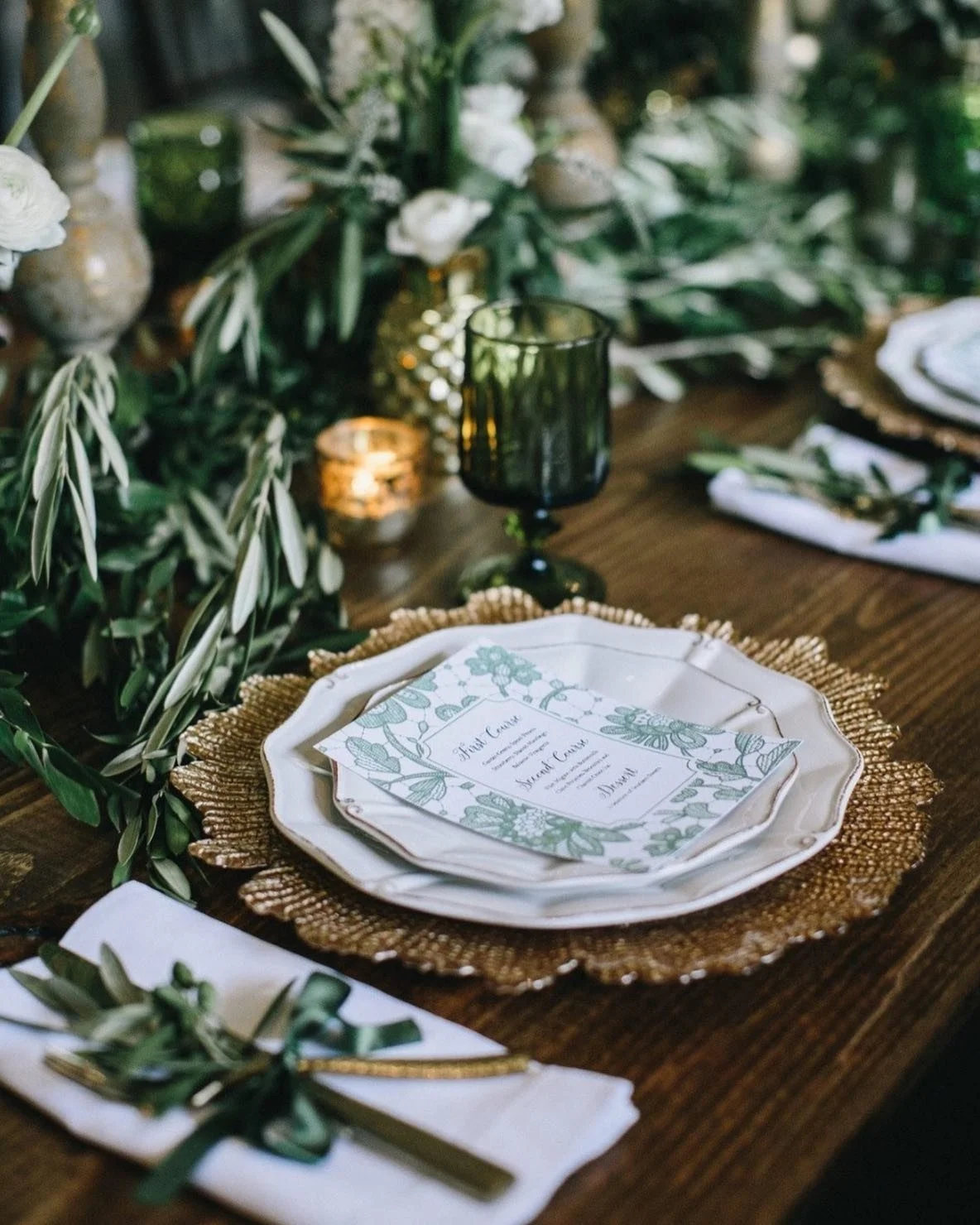 Elegant wedding table setting with ornate plates, leafy green and white floral centerpiece, gold accents, candlelight, and a menu card.