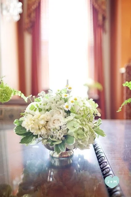 A white floral centerpiece on a polished wooden table with red curtains and a window in the background.