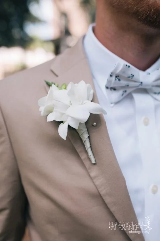 Close-up of a man wearing a beige suit, white shirt, and a bow tie, with a white boutonniere pinned to his lapel.