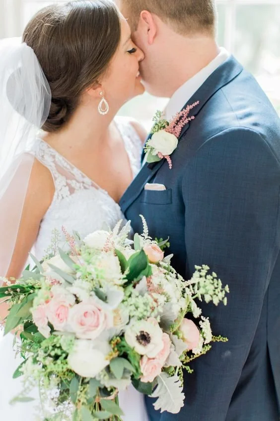 A bride and groom sharing a kiss on their wedding day, with the bride holding a large pastel-colored bouquet and both dressed in wedding attire.