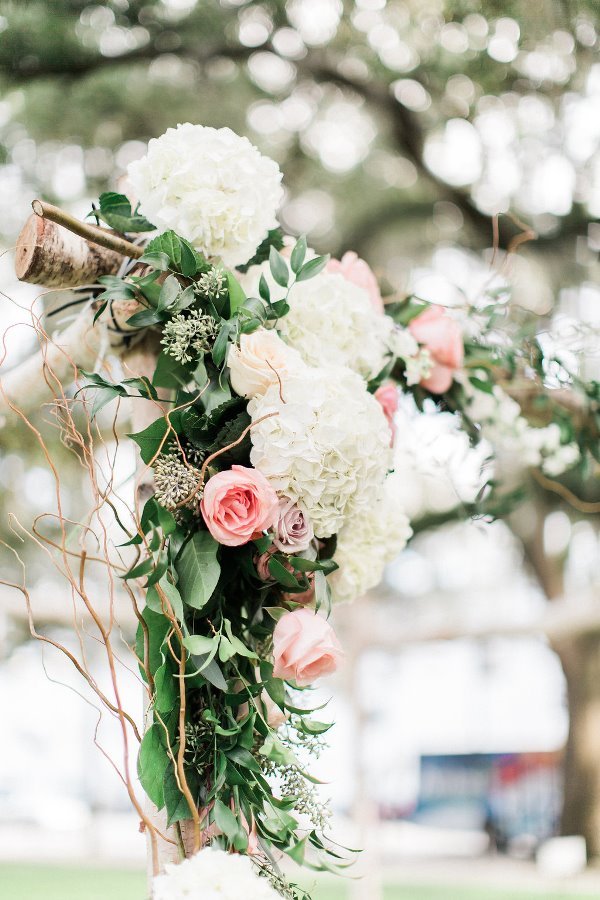 Decorative floral arrangement with white hydrangeas, pink roses, and green foliage, hanging outdoors with blurred trees and cars in the background.