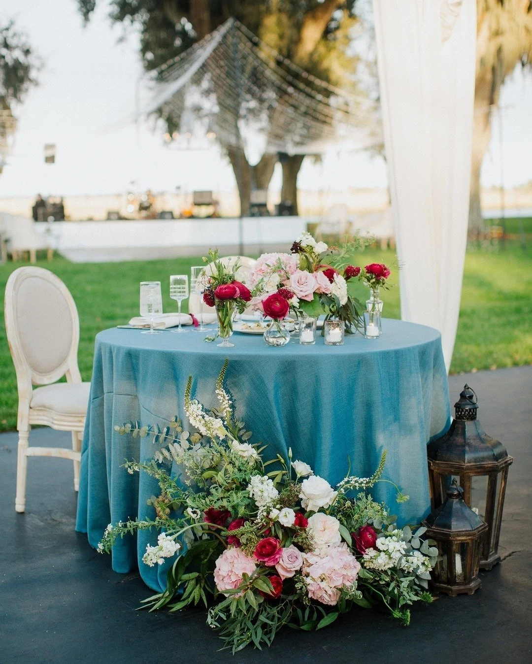 Decorated outdoor table with blue tablecloth, floral centerpieces, candles, and lanterns, set for an event with chairs and trees in the background.