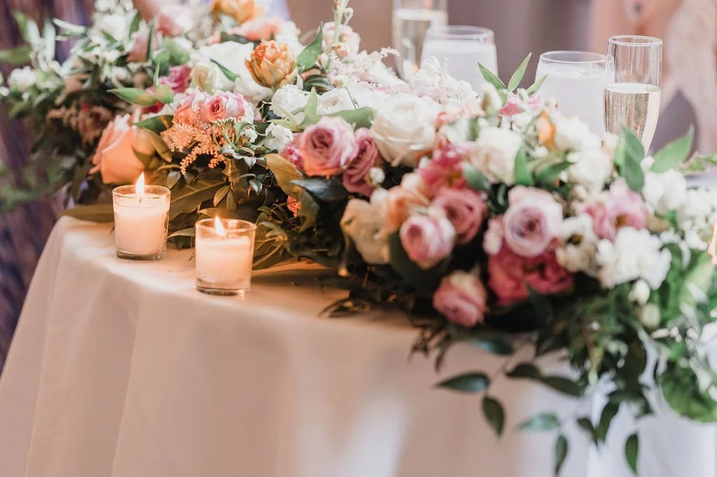 Elegant table decorated with a floral centerpiece of pink and white roses and greenery, with lit candles and glasses of water.