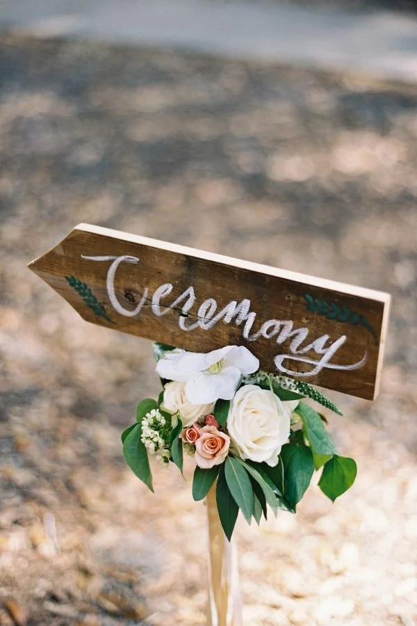 Wooden wedding sign with white and pink roses and green leaves, reading 'Ceremony' in white script, outdoors on dirt ground.