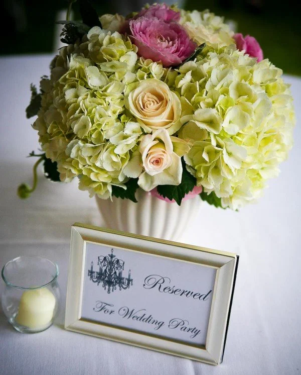 A floral arrangement with white hydrangeas, pink roses, and cream roses in a white vase, placed on a table with a "Reserved for Wedding Party" sign and a candle in a glass holder.