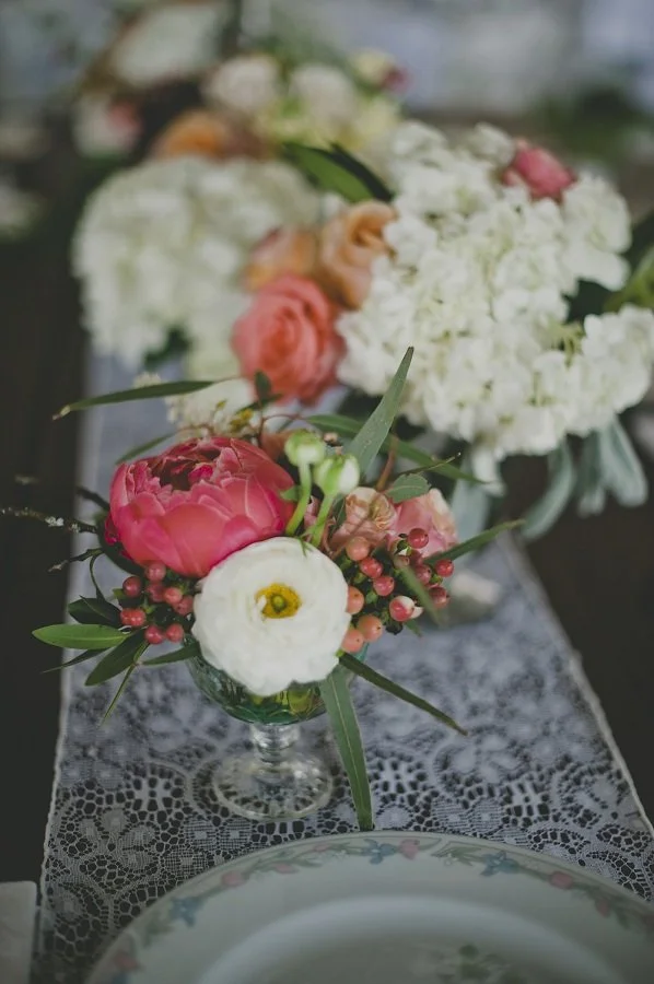 Close-up of a floral centerpiece with pink peony, white ranunculus, pink hypericum berries, and green leaves on a lace tablecloth with blurred bouquet in background.
