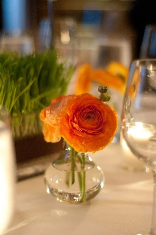 A small glass vase with an orange ranunculus flower on a table, with a salad and glassware in the background.