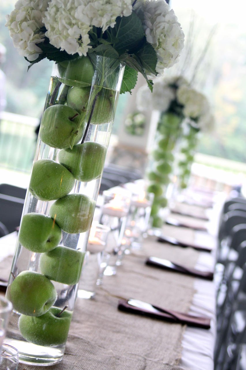A tall glass vase filled with water and green apples, topped with white hydrangea flowers. The vase is part of a decorated table with white chairs and elegant place settings, set up for a gathering or event.