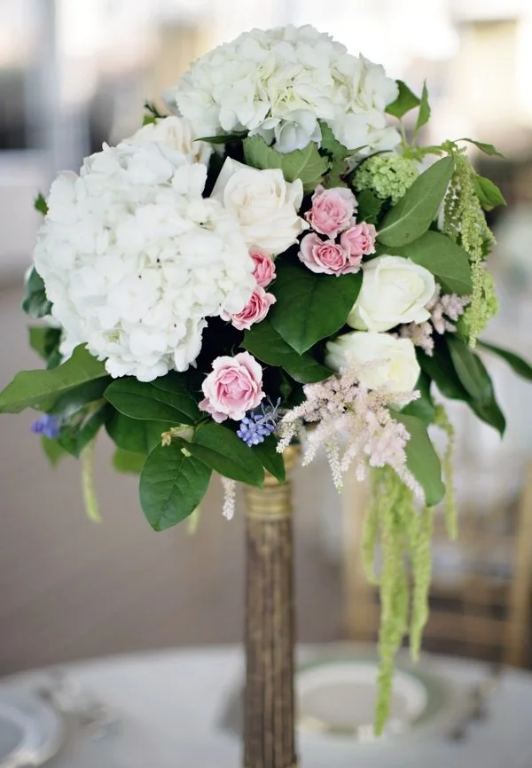 A bouquet of white hydrangeas, roses, and greenery in a tall wooden stand.