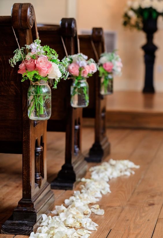 Wooden church pews decorated with glass jars holding pink and white flowers, with white flower petals scattered on the wooden floor in an indoor setting.