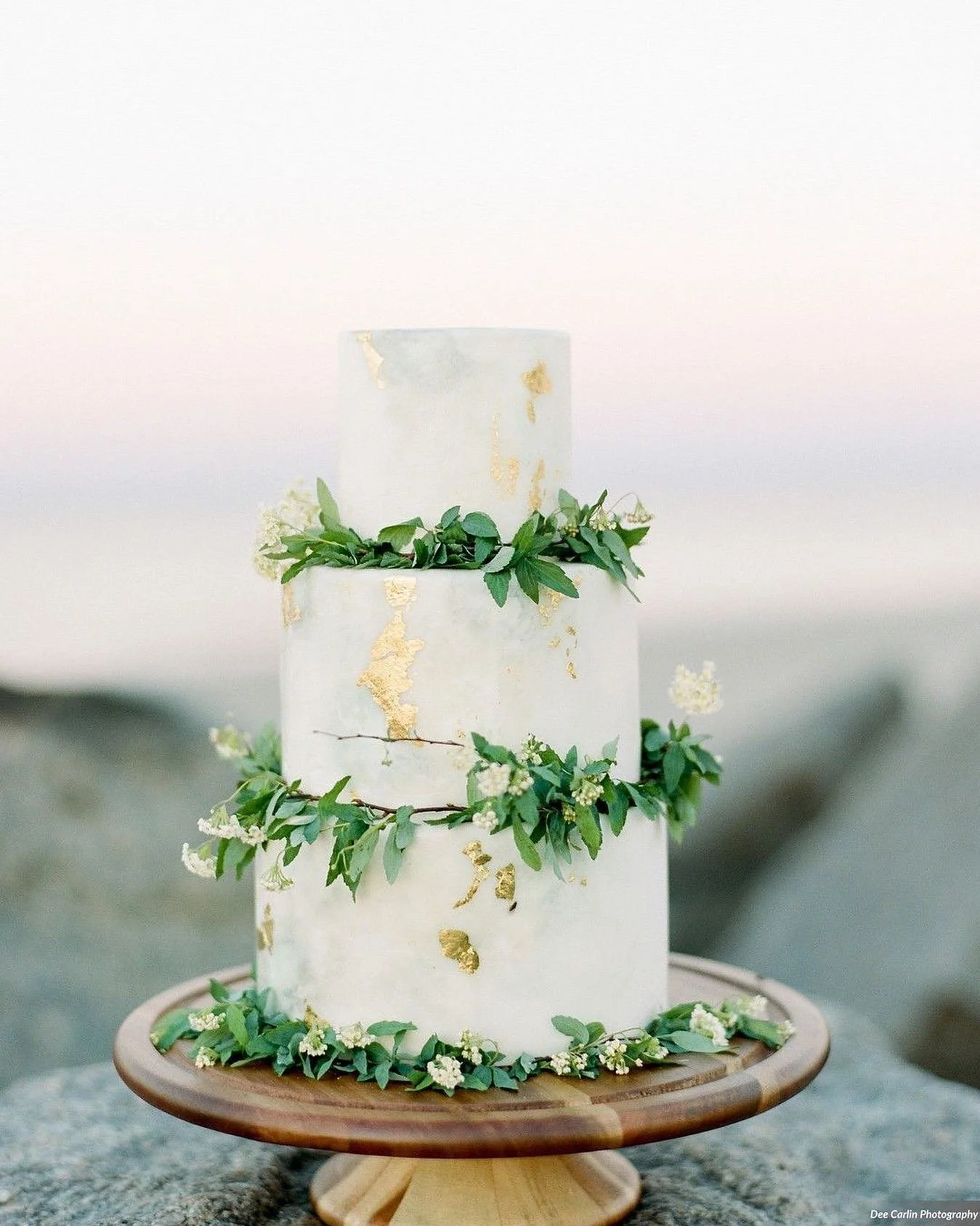 Three-tiered white wedding cake with gold leaf accents, decorated with greenery and small white flowers, placed on a wooden cake stand outdoors.