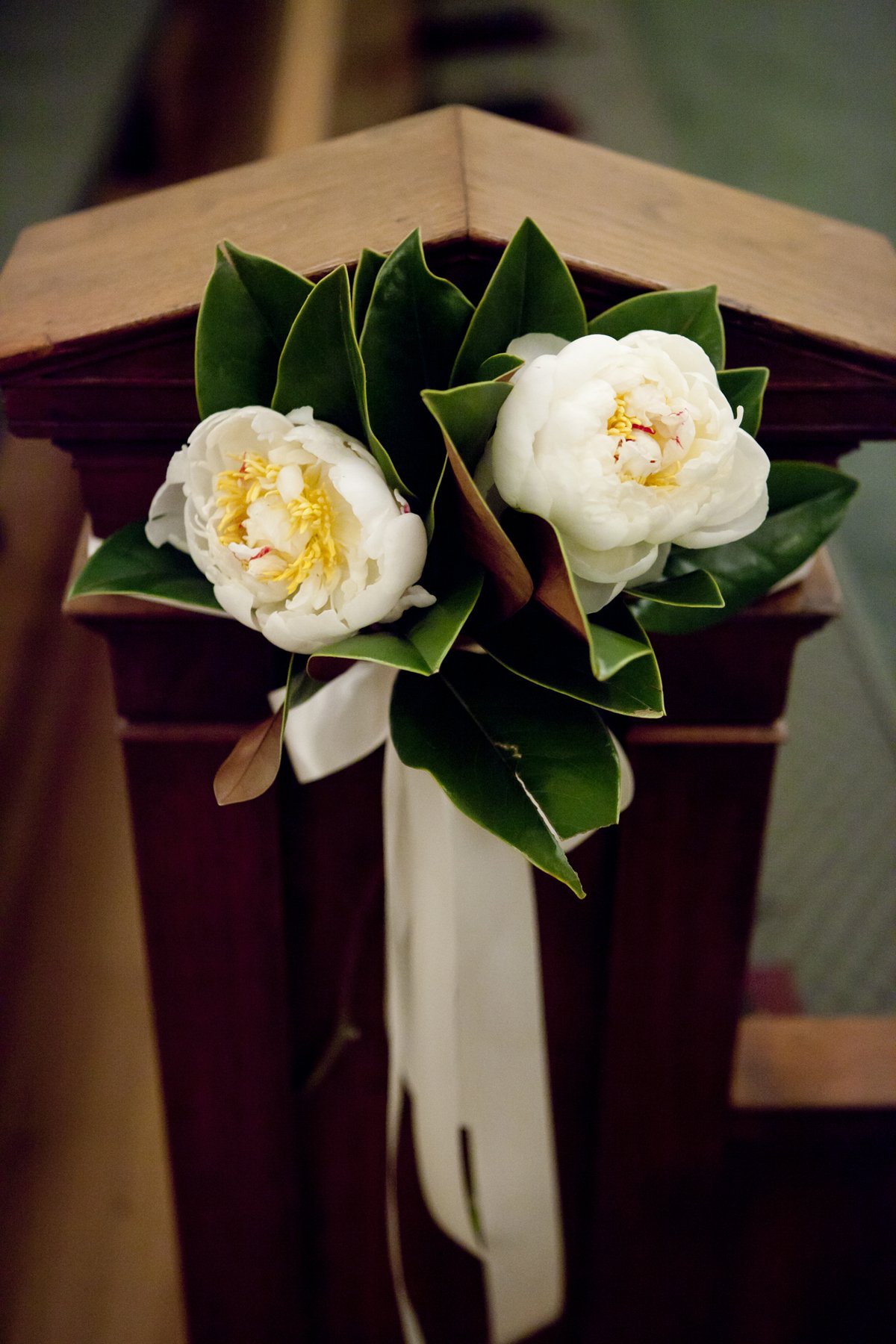 A small bouquet of white flowers with yellow centers, surrounded by dark green leaves, attached to a ribbon, resting on a wooden surface.