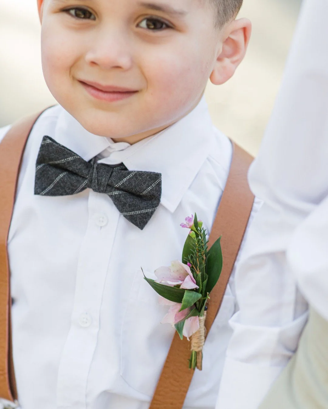 Close-up of a young boy dressed formally with a white shirt, a grey checkered bow tie, brown suspenders, and a boutonniere pinned to his shirt, smiling outdoors.