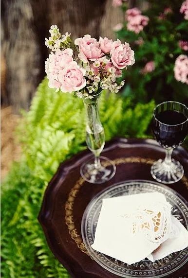 A table with a glass of red wine, a decorative white napkin, and a small bouquet of pink and white flowers in a tall glass vase, set outdoors on a brown tray.